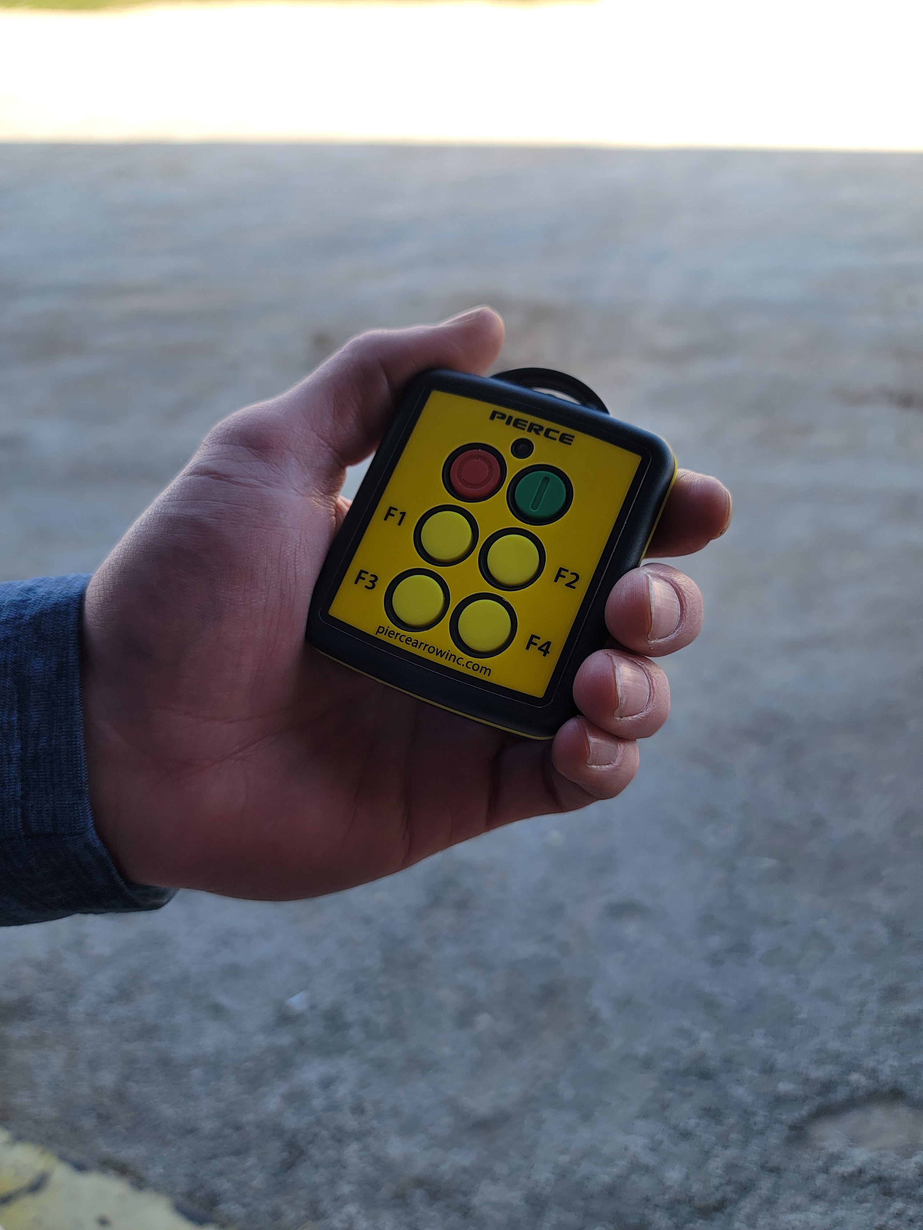 A hand holds a PIERCE 4 Function Wireless Remote Transmitter with six buttons labeled F1 to F4, plus green and red buttons. This sleek device offers seamless fingertip control and acts as a replacement transmitter.