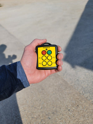 In an outdoor setting, a hand holds a yellow PIERCE 4 Function Wireless Remote Transmitter with black edges and six buttons labeled F1 to F4.