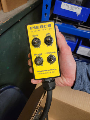 A hand holds a yellow PIERCE 8 Function Wrecker Remote with four switches labeled for boom, jaws, and folding. Ideal for towing, this wired remote integrates smoothly with advanced solenoid systems for optimal performance.