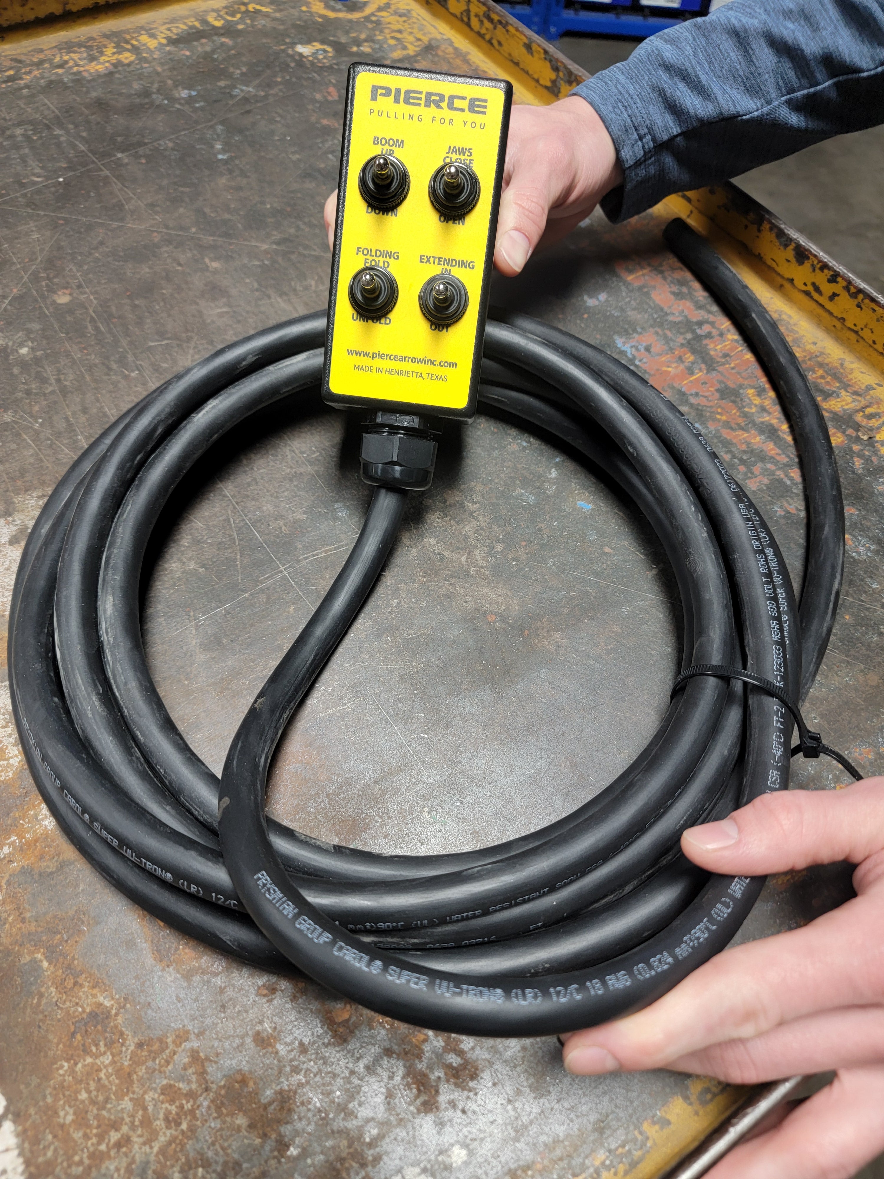 A person holds a black coiled cable with a yellow PIERCE control box. It features the 8 Function Wrecker Remote, perfect for solenoid systems in towing, and includes an intuitive toggle switch wired remote for seamless operation.