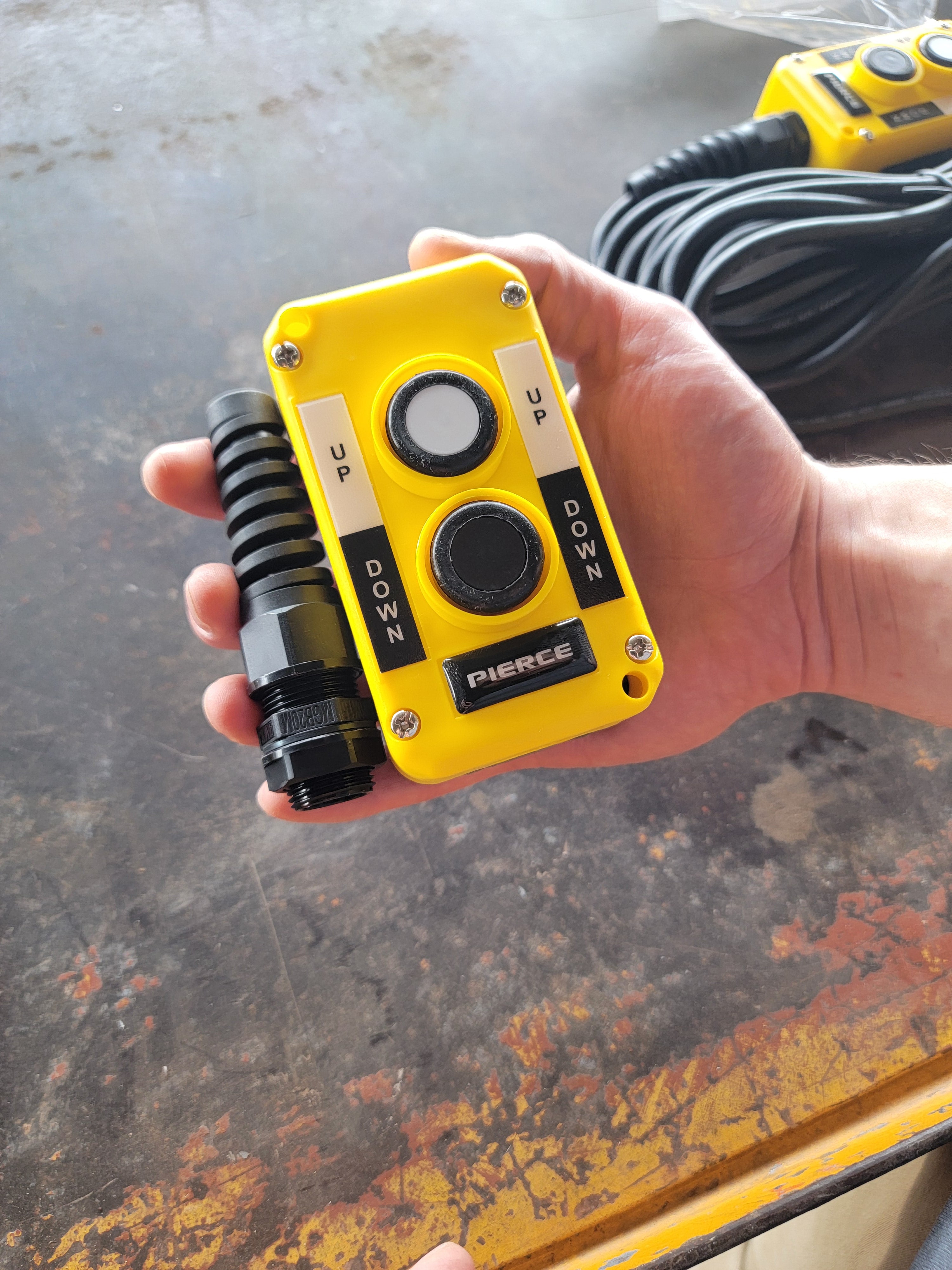 A hand grips a PIERCE Universal Momentary Pendant Control, featuring UP and DOWN buttons on a yellow design with a coiled cable. Perfect for industrial use, it ensures smooth operation of hydraulic power units.