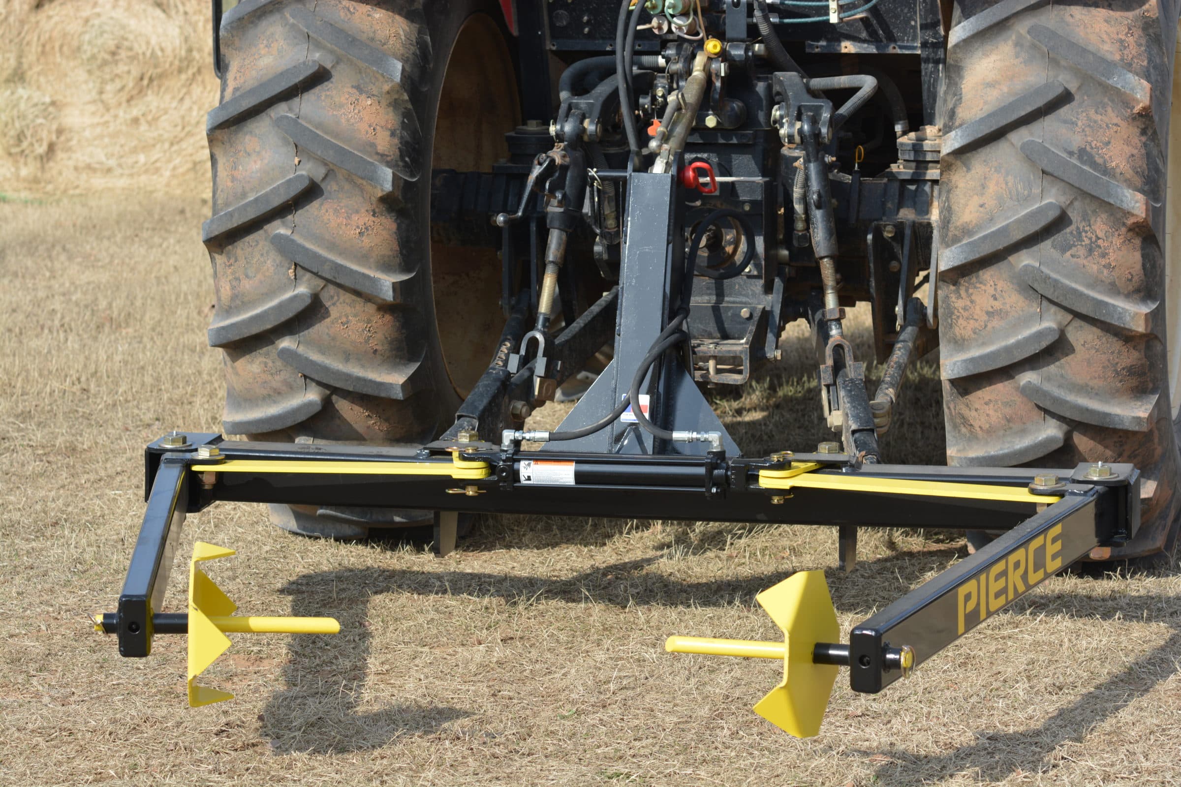 A PIERCE 3-Point Hay Bale Unroller with a black and yellow frame is set on the dry grassy field, prepared to handle large round bales effortlessly.