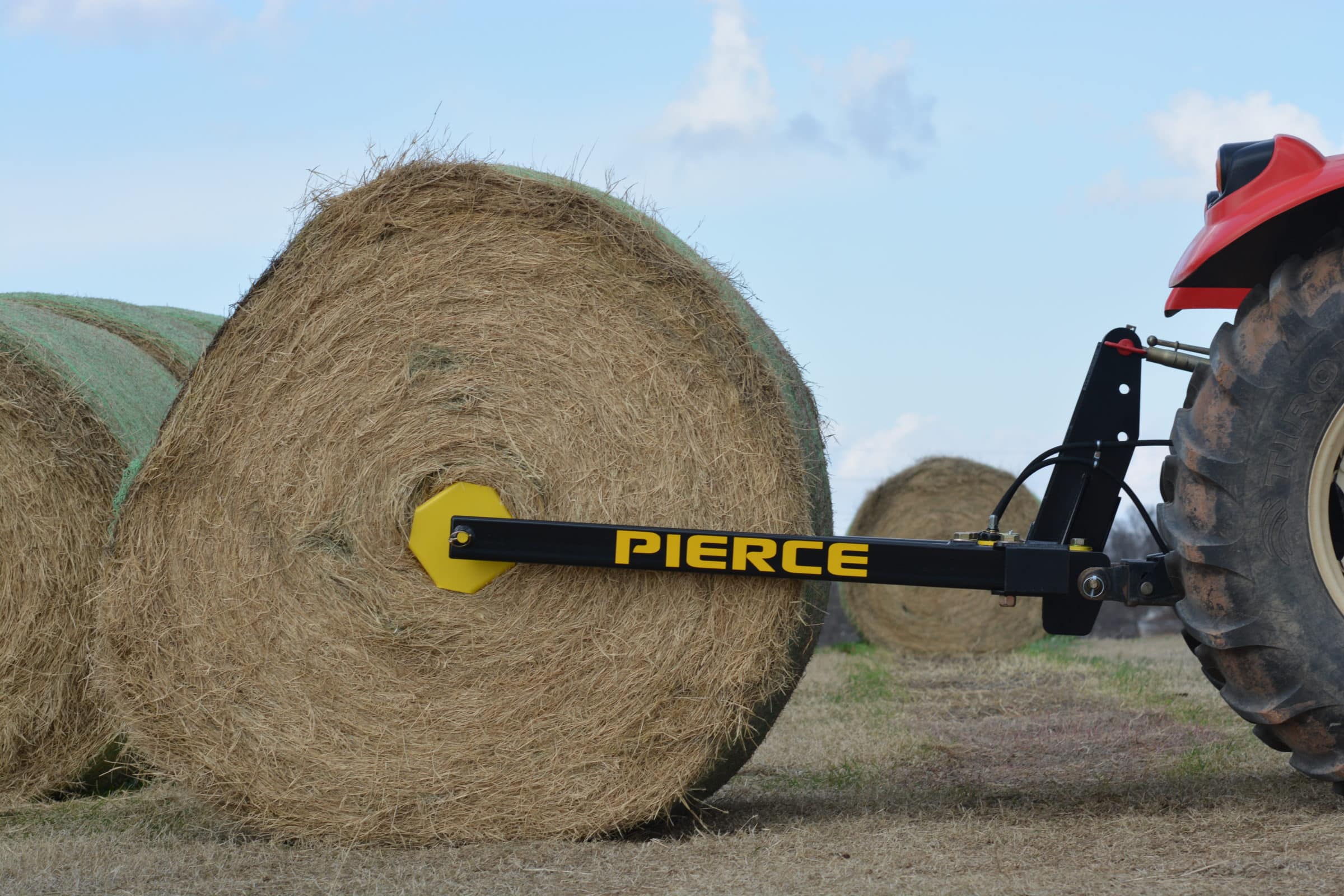 A tractor navigates the field under a blue sky, skillfully moving large round bales with its PIERCE 3-Point Hay Bale Unroller.