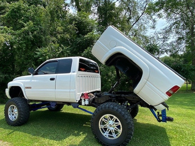 A lifted white pickup truck equipped with a PIERCE Factory Bed Dump Kit, featuring a hydraulic hoist for the tilt bed, is parked on a grassy area surrounded by trees.