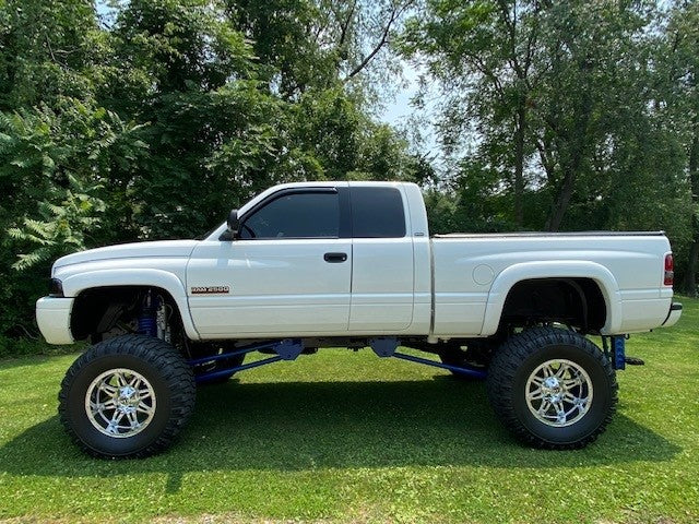 A white pickup truck, enhanced with the PIERCE Factory Bed Dump Kit, sits majestically on large tires parked on grass, surrounded by trees in the background.