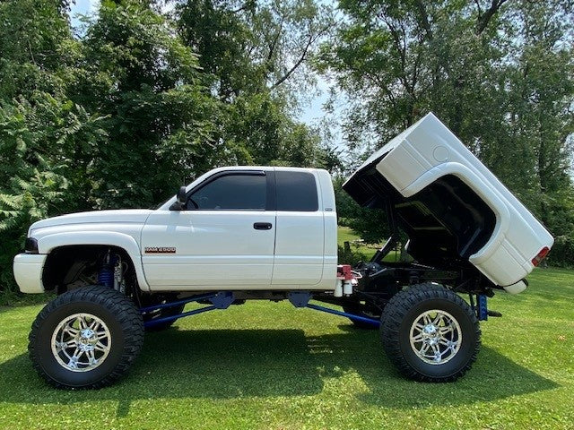 A white truck equipped with the PIERCE Factory Bed Dump Kit stands on grassy terrain, its bed tilted open, surrounded by leafy trees.