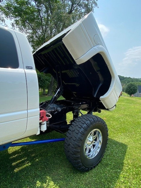 A truck equipped with a PIERCE Factory Bed Dump Kit, featuring lifted cab exposing the engine, large off-road tires, and a powerful hydraulic hoist kit, is parked on grass with trees in the background.