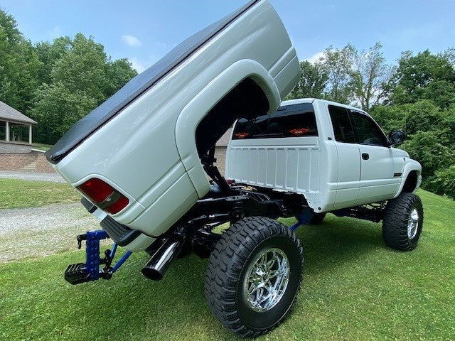 A lifted white pickup truck equipped with PIERCE's Factory Bed Dump Kit, featuring a hydraulic hoist and tilting bed, stands proudly on a grassy area near trees and a small building, showcasing its impressive lifting strength.