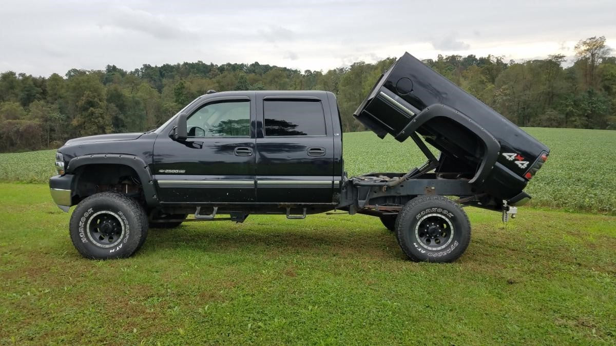 A truck with a lifted suspension and a tilted bed, equipped with the PIERCE Factory Bed Dump Kit hydraulic hoist, rests in a grassy field surrounded by trees.