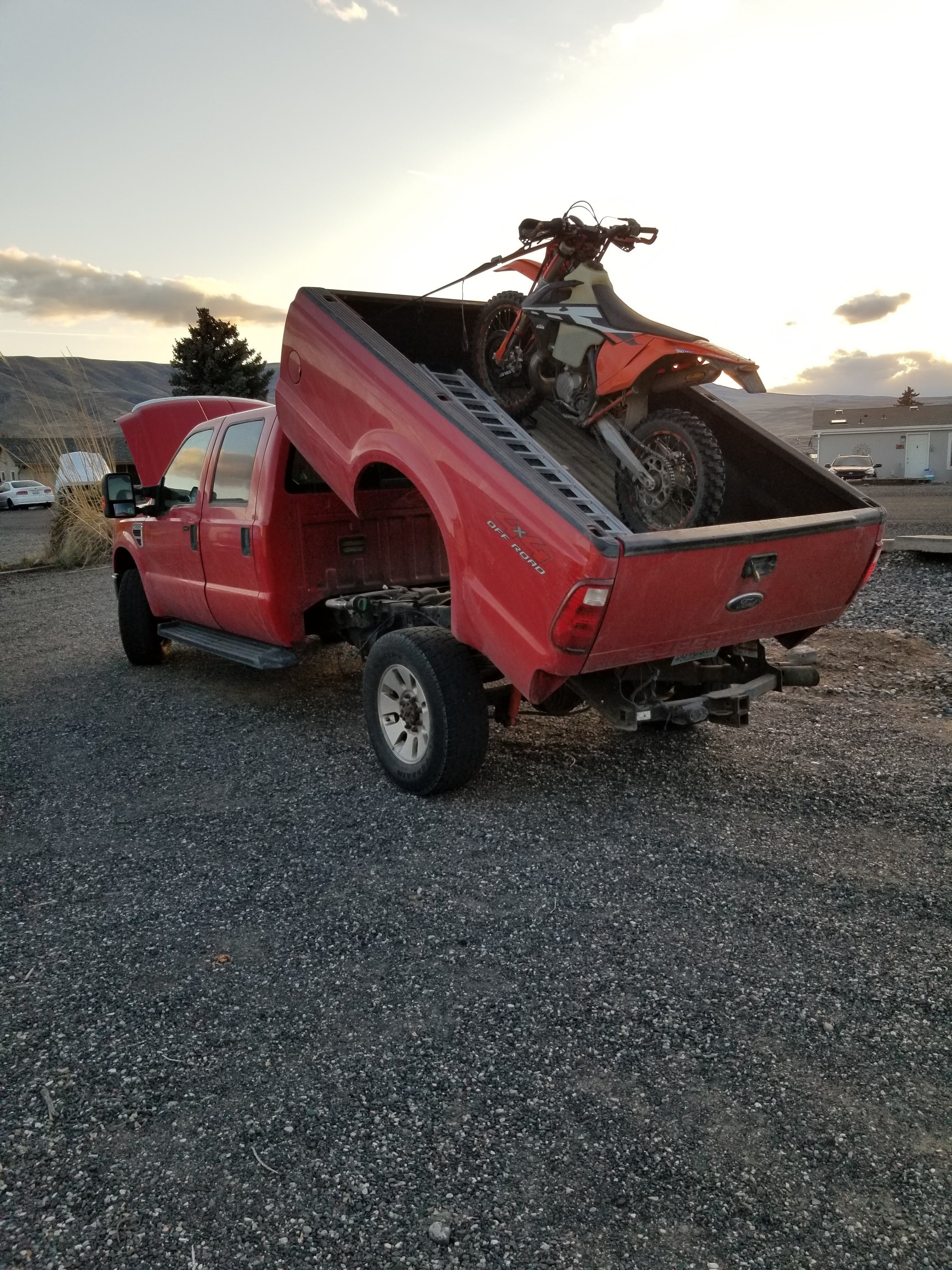 A PIERCE Factory Bed Dump Kit-equipped red truck stands upright on a gravel lot at sunset, demonstrating its impressive lifting strength by effortlessly holding a motorcycle.